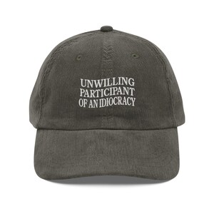 May include: A dark green corduroy baseball cap with white embroidered text that reads "Unwilling Participant of an Idiocracy".