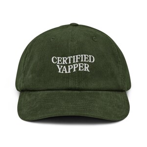 May include: A green corduroy baseball cap with the text "CERTIFIED YAPPER" embroidered in white. The cap has a curved brim and an adjustable strap at the back. The hat is a casual accessory.