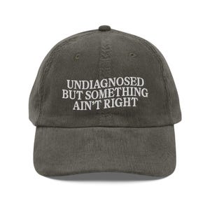 May include: A dark green corduroy baseball cap with white embroidered text that reads "Undiagnosed but something ain't right".