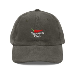 Puede incluir: Gorra de béisbol de pana verde oliva. Presenta un logotipo de pimiento rojo con tallo verde y el texto blanco "Neurospicy Club". La gorra tiene una visera curva y una correa ajustable.