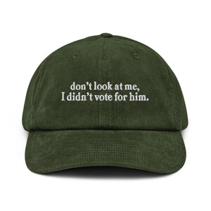 以下が含まれることがあります： カーキ色のコーデュロイベースボールキャップ。白い刺繍で「don't look at me, I didn't vote for him」と書かれています。キャップはカーブしたつばと、後ろに調節可能なストラップが付いています。