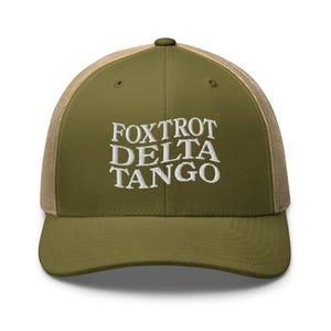 以下が含まれることがあります： 前面に白い刺繍で「FOXTROT DELTA TANGO」と書かれた、緑とベージュのトラッカーハットです。