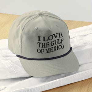 May include: A beige trucker hat with a black brim and a black embroidered message that reads "I Love The Gulf Of Mexico".
