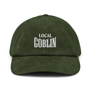 May include: A dark green corduroy baseball cap with the embroidered text "LOCAL GOBLIN" in white. The cap has a curved brim and a structured crown. The hat is a casual accessory.