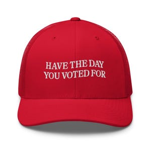 Op de afbeelding: Rode mesh trucker cap met witte geborduurde tekst die "Have the day you voted for" luidt.