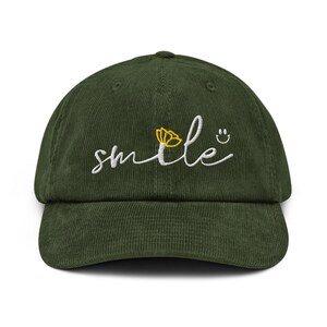May include: A dark green corduroy baseball cap. The cap features the word "smile" embroidered in white script, a yellow flower, and a smiling face. The cap has a curved peak.