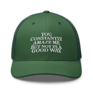 May include: Green trucker hat with the text "YOU CONSTANTLY AMAZE ME BUT NOT IN A GOOD WAY" embroidered in white. The hat has a mesh back and a curved brim.