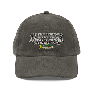 Puede incluir: Una gorra de béisbol de pana verde oscuro con bordado blanco que dice "Let the fish who thinks he knows no fear look well upon my face". Una pequeña imagen bordada de un salmón está debajo del texto.
