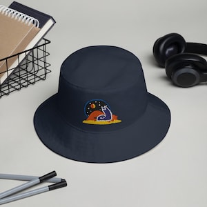 May include: A navy blue bucket hat with an embroidered design of a howling wolf under a starry night sky. The design includes a red sun and yellow ground. The hat is a casual accessory.