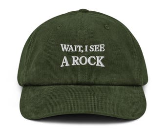 Puede incluir: Gorra de béisbol de pana verde oliva con la frase bordada "WAIT, I SEE A ROCK" en blanco. La gorra tiene una visera curva y una corona estructurada. Un accesorio casual.