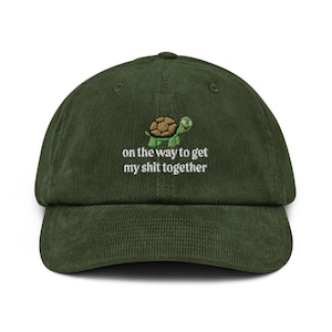May include: A green corduroy baseball cap with a curved peak. The cap has an embroidered cartoon turtle and the phrase "on the way to get my shit together" in white. The cap is a solid colour.