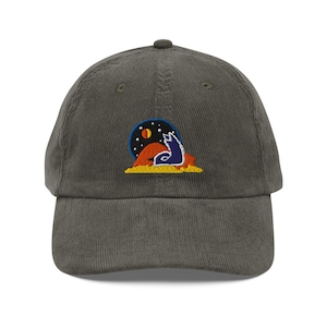 May include: A corduroy baseball cap in a muted green color. The cap features an embroidered patch with a blue wolf howling at a moon and stars over a desert landscape. The design is outlined in yellow and set against a dark blue background.
