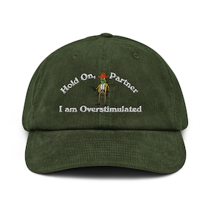 Puede incluir: Gorra de béisbol de pana verde oscuro con la frase bordada "Hold On, Partner, I am Overstimulated". La gorra presenta una rana de dibujos animados bordada con sombrero de vaquero y chaleco.