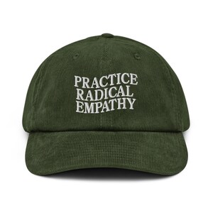 May include: A dark green corduroy baseball cap. The front features the embroidered text "PRACTICE RADICAL EMPATHY" in white. The cap has a curved peak and an adjustable strap at the back.