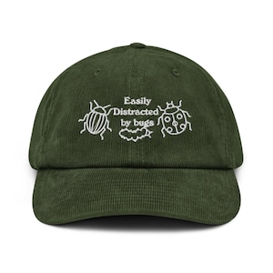 May include: Dark olive green corduroy baseball cap with white embroidered text and bug illustrations. The text reads "Easily Distracted by bugs". The cap has a curved peak and an adjustable strap at the back.