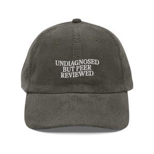 May include: An olive green corduroy baseball cap featuring the embroidered text "UNDIAGNOSED BUT PEER REVIEWED" in white. The cap has a curved peak and a rounded crown. A casual hat.