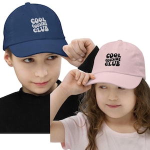May include: Two baseball caps, one navy blue and one light pink, each with the text "COOL COUSINS CLUB" in a white, bubble-style font. The caps are being held up by children.