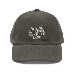 May include: A dark green corduroy baseball cap with white embroidered text that reads "NO ONE IS ILLEGAL ON STOLEN LAND".