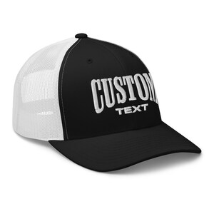 May include: Black and white trucker hat with a white mesh back. The front of the hat has white embroidered text that says "CUSTOM TEXT".