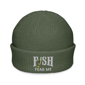 Könnte beinhalten: Olivgrüne Mütze mit dem gestickten Text "FISH FEAR ME" in Weiß. Das Wort "FISH" hat eine farbenfrohe Fischgrafik. Die Mütze ist aus einem Rippstrickmaterial gefertigt.