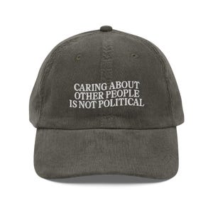 May include: Olive green corduroy baseball cap with the embroidered text: "CARING ABOUT OTHER PEOPLE IS NOT POLITICAL". The cap has a curved brim and a fabric-covered button on top. A casual accessory.