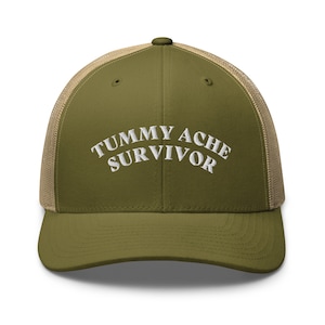 May include: A green and tan trucker hat with the text "TUMMY ACHESURVIVOR" embroidered in white.