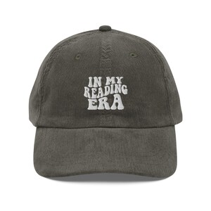 Op de afbeelding: Olijfgroene corduroy baseballcap met de geborduurde witte tekst "IN MY READING ERA". De cap heeft een gebogen klep en een afgeronde bovenkant, geschikt voor casual wear.