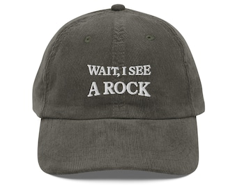 Puede incluir: Gorra de béisbol de pana verde oliva con el texto bordado "WAIT, I SEE A ROCK" en blanco. La gorra tiene una visera curva y un botón en la parte superior.