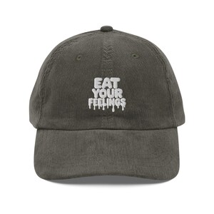 Könnte beinhalten: Dunkel olivgrüne Cord-Baseballkappe mit gebogenem Schirm. Auf der Kappe steht in weißer Schrift "EAT YOUR FEELINGS" in einem tropfenden Schriftzug. Die Kappe hat eine runde Krone und einen kleinen Knopf oben.