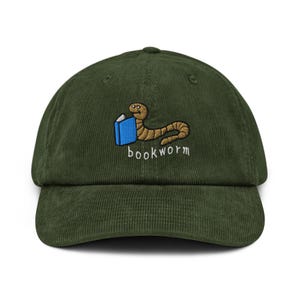 May include: A dark green corduroy baseball cap with an embroidered design of a cartoon bookworm reading a blue book. The word "bookworm" is embroidered below the graphic in white.