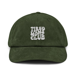 May include: A dark green corduroy baseball cap with the text "TIRED MOMS CLUB" embroidered in white. The cap has a curved brim and a structured crown, suitable for casual wear.