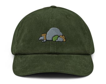Puede incluir: Gorra de béisbol de pana verde oscuro con visera curva. La gorra presenta un diseño bordado de una roca gris con detalles en verde y marrón. Un accesorio clásico e informal.