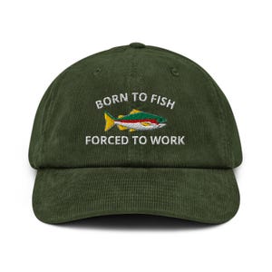 以下が含まれることがあります： 濃い緑色のコーデュロイ野球帽。刺繍された「BORN TO FISH FORCED TO WORK」の文字と、カラフルな魚のグラフィックが特徴です。魚は黄色、赤、緑、白のディテールがあります。帽子はカーブしたつばと構造化されたクラウンを持っています。