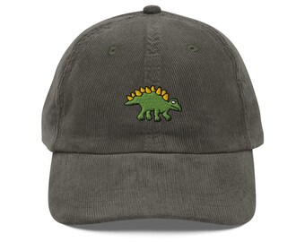 May include: A corduroy baseball cap in a muted olive green color. The cap features a small embroidered dinosaur in green with yellow spines on the front. The cap has a curved brim and a fabric strap closure.