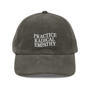 Może przedstawiać: Oliwkowozielona czapka bejsbolówka z sztruksu z białym haftem "PRACTICE RADICAL EMPATHY". Czapka ma zakrzywiony daszek i regulowany pasek. Casualny dodatek do noszenia na głowie.