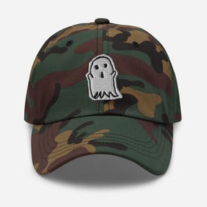 Puede incluir: Una gorra de béisbol de camuflaje con un diseño de fantasma bordado en blanco.