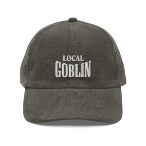 May include: A dark green corduroy baseball cap with white embroidered text that reads "LOCAL GOBLIN".