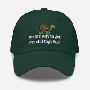 May include: A dark green baseball cap with an embroidered image of a turtle and the text "on the way to get my shit together".