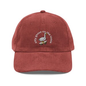 Puede incluir: Gorra de béisbol de pana color ladrillo con visera curva. La gorra presenta un diseño bordado de un ganso en monopatín, con el texto "silly goose on the loose" en arco circular.