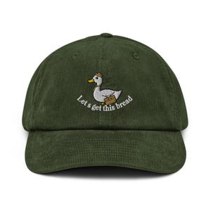 Può includere: Cappellino da baseball in velluto a coste verde scuro con visiera curva. Il cappellino presenta un ricamo di un'anatra con cappello e valigetta, con la scritta "Let's get this bread" in bianco.