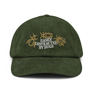 Puede incluir: Gorra de béisbol de pana verde oscuro con la frase bordada en dorado "EASILY DISTRACTED BY BUGS". El diseño incluye ilustraciones estilizadas de insectos. La gorra tiene una visera curva y una corona estructurada.