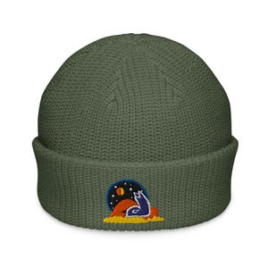 May include: Olive green beanie with a folded brim. The beanie features an embroidered patch with a blue wolf howling at a moon and stars over a desert landscape. The beanie is made of a ribbed knit material.
