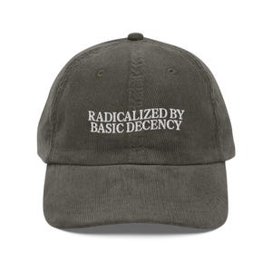 May include: Olive green corduroy baseball cap with the text "RADICALIZED BY BASIC DECENCY" embroidered in white. The cap has a curved brim and a small button on top. A casual accessory.