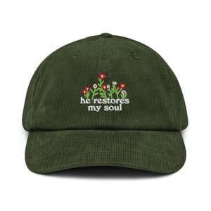 Op de afbeelding: Donkergroene corduroy baseballcap met een gebogen klep. De cap heeft een geborduurd bloemendesign in rood en wit, met de tekst "he restores my soul" in witte letters. Een klassiek, casual accessoire.