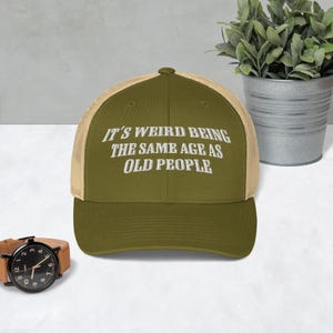 May include: A green and beige trucker hat with the text "It's weird being the same age as old people" embroidered in white.