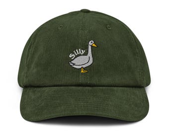 May include: A dark green corduroy baseball cap with a curved brim. The cap features an embroidered design of a gray goose with the word "Silly" above it. The goose has a yellow beak and feet.