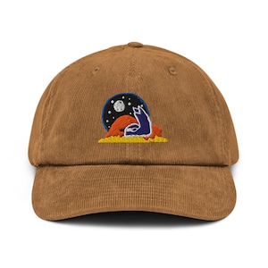 May include: A brown corduroy baseball cap with an embroidered design. The design features a blue wolf howling at a moon and stars, set against a dark blue background. The wolf is on a yellow and orange landscape.