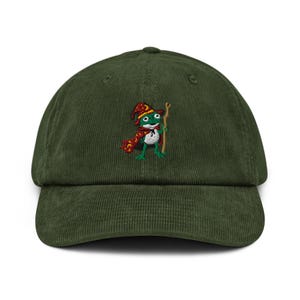 May include: Green corduroy baseball cap with an embroidered cartoon frog wizard. The frog wears a red and gold hat and scarf, and holds a wooden staff. The cap has a curved peak and an adjustable strap at the back.