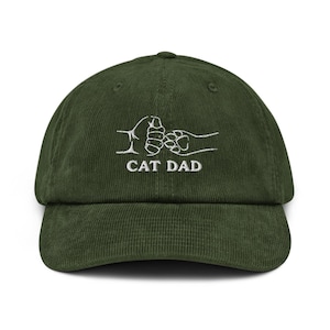 May include: A green corduroy baseball cap with a curved peak. The cap has white embroidered text and a graphic of a human hand bumping a cat paw. The text reads "CAT DAD".