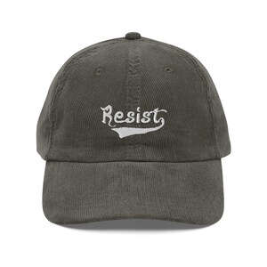 Könnte beinhalten: Eine dunkelgrüne Baseballkappe aus Cord mit weiß gesticktem Text, der "Resist" in einer Vintage-Schriftart lautet.